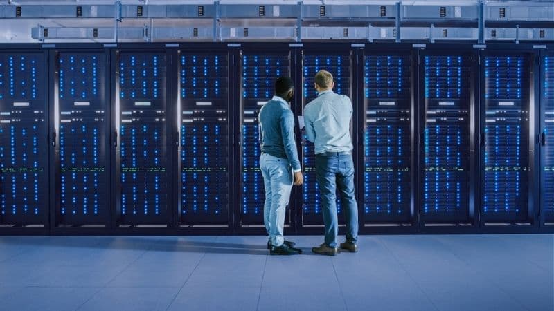 Bearded IT Technician In Glasses With A Laptop Computer And Black Male Engineer Colleague Are Talking In Data Center
